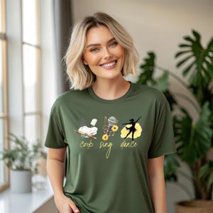 Puede incluir: Una camiseta verde con un diseño gráfico de un rodillo, un micrófono y una bailarina. El texto "cook sing dance" está escrito debajo del gráfico.