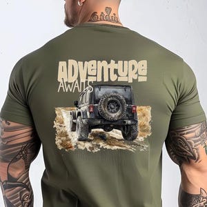 Op de afbeelding: Een groen T-shirt met een afbeelding van een jeep die over een onverharde weg rijdt. De tekst 'Adventure Awaits' staat boven de jeep gedrukt.