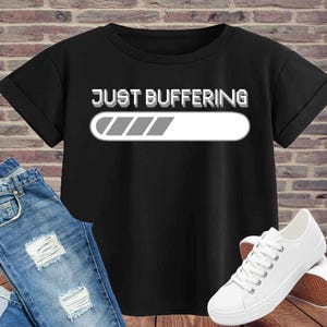 Puede incluir: Camiseta negra con el texto "JUST BUFFERING" y una barra de carga. Se combina con vaqueros azules y zapatillas de lona blancas. Un conjunto casual y moderno.