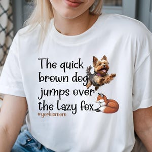 May include: White t-shirt featuring a graphic of a Yorkshire Terrier dog jumping over a fox, with the text "The quick brown dog jumps over the lazy fox." The hashtag #yorkiemom is also included.