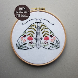Peut inclure: Broderie de papillon de nuit dans un cercle de broderie en bois. Le papillon de nuit est blanc avec des feuilles vertes et des fleurs roses sur ses ailes.