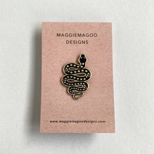May include: Black and gold enamel pin featuring a stylised snake with a dotted pattern. The pin is on a brown card with the text "MAGGIEMAGOO DESIGNS" and "www.maggiemagoodesigns.com".