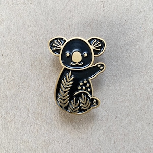 May include: Black and gold enamel pin featuring a koala with a floral design. The koala is standing on its hind legs with its arms raised.