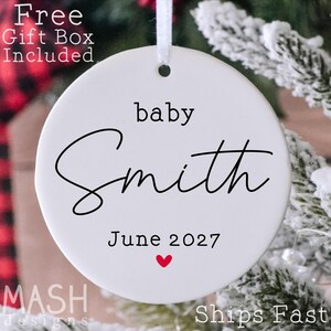 May include: A round white ceramic ornament featuring the black printed text baby Smith and June 2027 with a small red heart icon below the date. The ornament has a hole at the top for a white ribbon hanger and a smooth, flat surface finish.