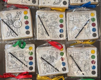 May include: Square-shaped cookies decorated with black and white line drawings of Toy Story characters, packaged in clear cellophane with colorful ribbons. Each cookie features a paint palette design with colored dots and a black edible marker.