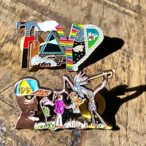 May include: Two enamel pins featuring artwork from the band Pink Floyd. The first pin is a colorful depiction of the band's name with various symbols. The second pin shows a man in a purple shirt standing next to a large, skeletal figure with a guitar.