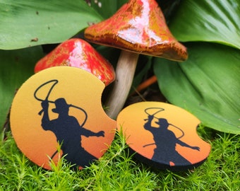 May include: Two round, yellow and orange coasters with a silhouette of a person holding a whip. The coasters are sitting on top of green moss.