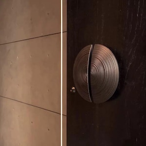 May include: A bronze-colored, circular door handle with a spiral design, split in two halves. The handle is mounted on a dark brown door, with a small, round doorknob visible. The background features a light-colored wall with horizontal lines.