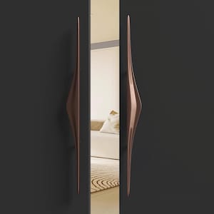 May include: Two bronze-colored, elongated door handles with a unique, curved design. The handles are mounted on a dark gray door, with a mirror reflecting a room interior. The handles have a sleek, modern aesthetic.