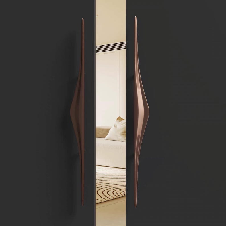 K&ouml;nnte beinhalten: Zwei l&auml;ngliche T&uuml;rgriffe in Bronzefarbe mit einem einzigartigen, geschwungenen Design. Die Griffe sind an einer dunkelgrauen T&uuml;r montiert, mit einem Spiegel, der einen Rauminnenraum reflektiert. Die Griffe haben eine elegante, moderne &Auml;sthetik.