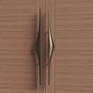 K&ouml;nnte beinhalten: Zwei bronzefarbene Schrankgriffe mit einem einzigartigen, l&auml;nglichen Tropfendesign. Die Griffe sind an einer Holzschrankt&uuml;r mit horizontalem Holzmaserungsmuster angebracht. Die Griffe haben eine glatte, reflektierende Oberfl&auml;che.