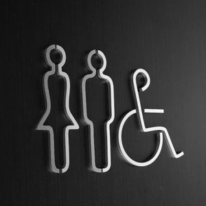 May include: A silver metal sign with three symbols: a female figure, a male figure, and a wheelchair. The symbols are outlined in silver and are on a black background.