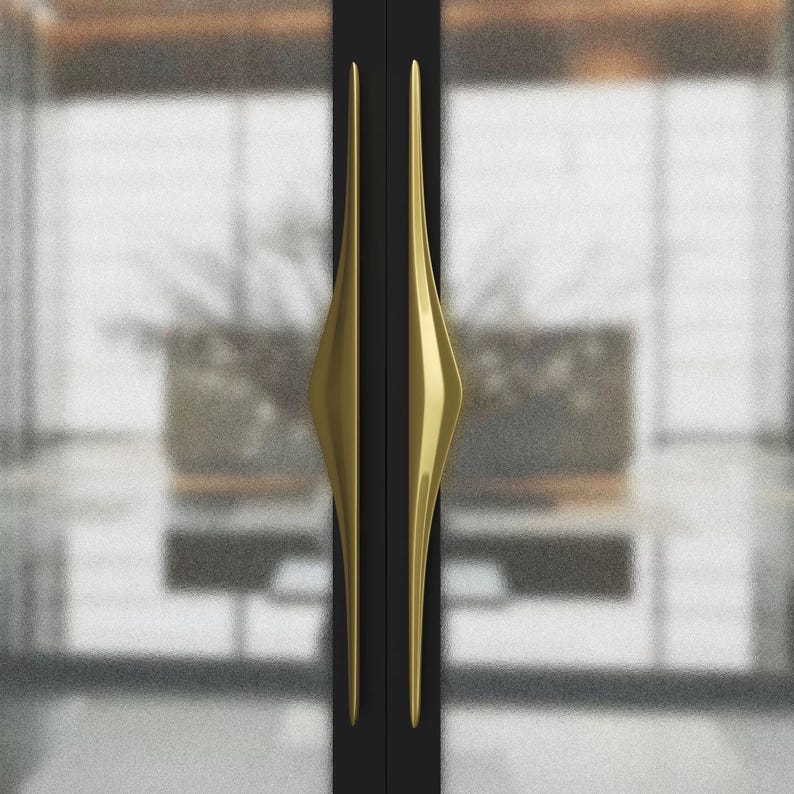 K&ouml;nnte beinhalten: Zwei schwarze T&uuml;ren mit goldfarbenen, l&auml;nglichen rautenf&ouml;rmigen Griffen. Die Griffe sind vertikal ausgerichtet und haben ein schlankes, modernes Design. Der Hintergrund ist verschwommen und zeigt einen Blick in einen Raum.