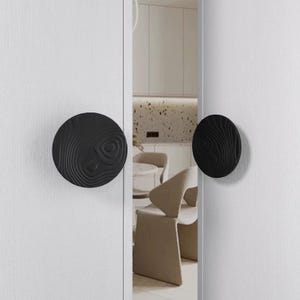 May include: Two black, circular door knobs with a wood grain design are mounted on a white door. The knobs are round with a textured surface, and the door is slightly ajar, revealing a glimpse of a modern interior.