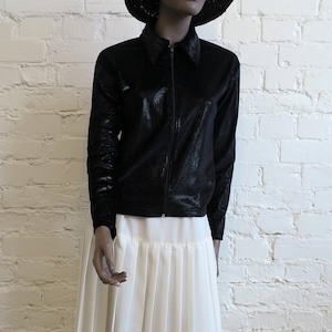 Può includere: Una giacca nera strutturata con chiusura a zip e colletto è abbinata a una gonna bianca a pieghe. L'outfit è esposto su un manichino contro un muro di mattoni bianchi e un pavimento in legno.