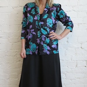 Op de afbeelding: Een zwarte blazer met een levendig bloemenpatroon in tinten blauwgroen en paars, gecombineerd met een effen zwarte midirok en kniehoge laarzen van zwart leer. De blazer heeft lange mouwen, een reverskraag en een knoopsluiting aan de voorzijde.