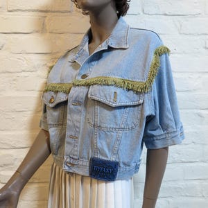 Puede incluir: Chaqueta vaquera azul claro con mangas cortas, flecos dorados y parche "Tiffany". La chaqueta tiene dos bolsillos en el pecho y dos bolsillos laterales. El maniquí lleva una falda plisada blanca.