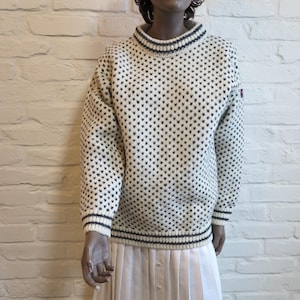 May include: Cream-colored sweater with a repeating black heart pattern. The sweater has a high, ribbed collar and cuffs with navy blue stripes. A small red and white patch is visible on the upper sleeve. The sweater is worn over a white pleated skirt.