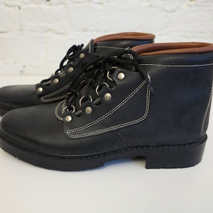 Könnte beinhalten: Ein Paar schwarze Lederstiefeletten mit braunem, gepolstertem Schaftrand, silberfarbenen Metallösen, schwarzen Schnürsenkeln und weißen Kontrastnähten an den Seitenteilen. Die Stiefel haben eine dicke schwarze Gummisohle und eine abgerundete Zehenpartie.