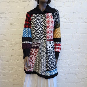 Op de afbeelding: Een lange patchwork trui met een zwarte kraag en manchetten. De trui heeft verschillende patronen in rood, wit, zwart, blauw en geel, waaronder geometrische vormen, bloemmotieven en dierenprints. De trui wordt op een mannequin gepresenteerd.