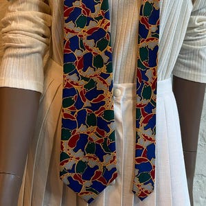 Peut inclure: Une cravate présentant un motif abstrait multicolore composé de formes rouges, bleues et vertes sur un fond beige clair. La cravate est portée sur un pull à col roulé blanc côtelé et une jupe plissée blanche, créant un ensemble superposé.