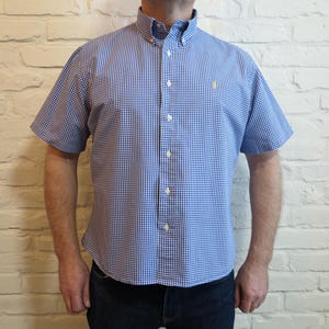 Könnte beinhalten: Kurzärmeliges Hemd mit blau-weißem Karomuster und Button-down-Kragen. Auf der Brust befindet sich ein kleines gelbes Emblem. Das Hemd wird mit einer dunkelblauen Jeans getragen. Klassisches Karomuster.