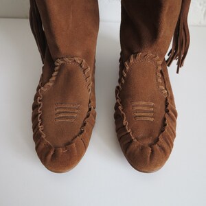 May include: A pair of brown suede boots with a moccasin-style design. The boots feature decorative stitching around the toe and a fringe detail at the top. The boots have a soft, textured appearance.