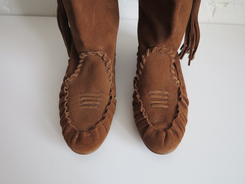 May include: A pair of brown suede boots with a moccasin-style design. The boots feature decorative stitching around the toe and a fringe detail at the top. The boots have a soft, textured appearance.