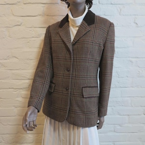 Pode incluir: Um blazer castanho aos quadrados com gola de veludo castanho escuro, dois botões frontais e dois bolsos com aba. O blazer é usado sobre uma camisola de gola alta branca e uma saia plissada branca, criando um visual estruturado em camadas.