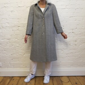 May include: A knee-length, grey plaid coat featuring a classic collar, long sleeves, and a front button closure. The coat is worn over a white shirt, white trousers, and white trainers, showcasing a structured fit with a subtle checkered pattern.