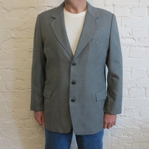 Könnte beinhalten: Ein grau-schwarzer Blazer mit Hahnentrittmuster und drei schwarzen Knöpfen. Der Blazer hat ein fallendes Revers und zwei Vordertaschen. Die Person trägt ein weißes Waffelstrickhemd und blaue Jeans.