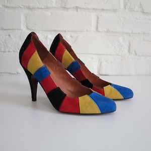 May include: A pair of high-heeled shoes with a colorful patchwork design. The shoes feature a combination of red, yellow, blue, and black suede panels. The heels are dark brown, and the shoes are set against a white background.