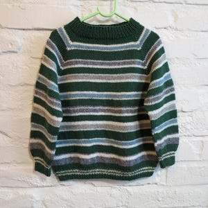 Può includere: Un maglione a righe verdi, bianche e blu con scollo tondo. Il maglione è appeso a un gancio verde. Le strisce orizzontali variano in larghezza. Il maglione è realizzato in maglia.