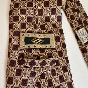 May include: A Joseph Abboud necktie featuring a repeating geometric pattern of dark maroon squares and tan diamond shapes. The back label reads Joseph Abboud, and a secondary tag indicates 100% seta-silk, made in Italy, fabrique en Italie.