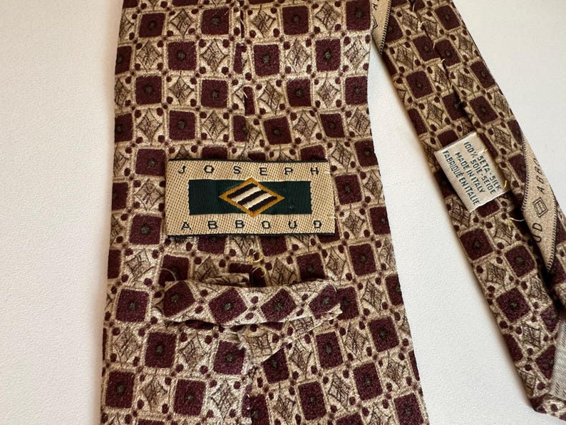May include: A Joseph Abboud necktie featuring a repeating geometric pattern of dark maroon squares and tan diamond shapes. The back label reads Joseph Abboud, and a secondary tag indicates 100% seta-silk, made in Italy, fabrique en Italie.