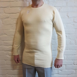 Könnte beinhalten: Cremefarbenes Langarm-Thermo-Shirt. Das Shirt hat einen Rundhalsausschnitt und eine enge Passform. Der Stoff scheint ein strukturiertes Strick zu sein. Die Ärmel enden in einem gerippten Bündchen. Das Shirt wird mit grauer Hose getragen.