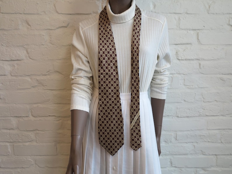 May include: A brown and tan patterned necktie featuring a repeating geometric motif. The tie is displayed against a white ribbed turtleneck sweater and a white pleated skirt, showcasing a classic design with a pointed tip and a fabric label.