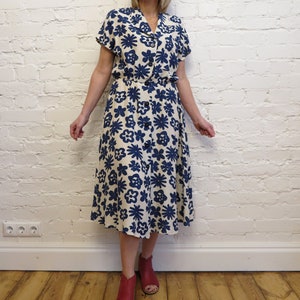 Puede incluir: Vestido color crema con estampado floral azul marino. Tiene mangas cortas, cuello camisero y cierre de botones. La falda plisada llega hasta la pantorrilla. El vestido se lleva con zapatos rojos.