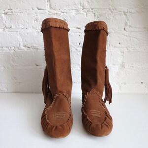May include: A pair of brown suede boots with a knee-high design. The boots feature a fringed top, decorative stitching, and a tassel detail. The footwear has a moccasin-style toe and a flat sole, suitable for casual wear.