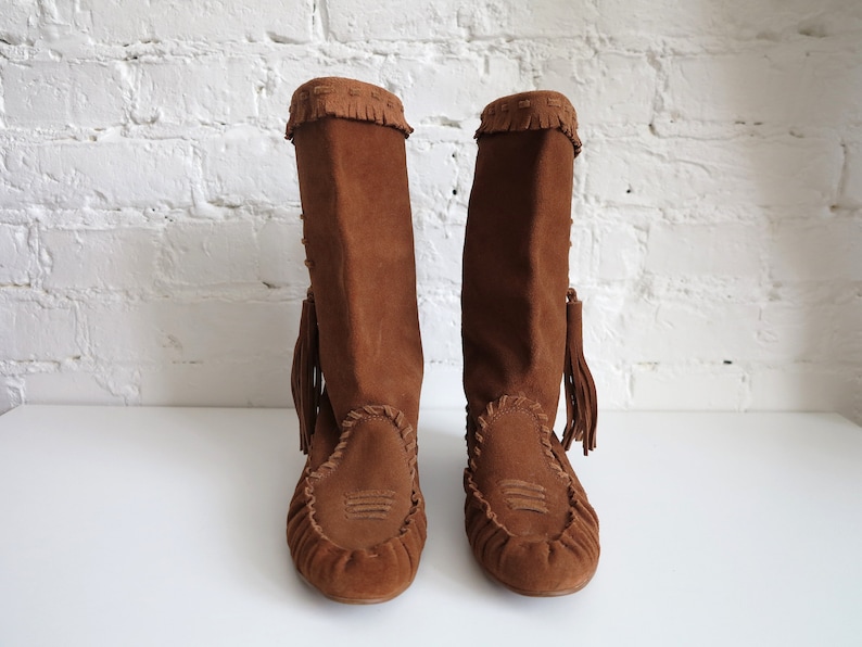 May include: A pair of brown suede boots with a knee-high design. The boots feature a fringed top, decorative stitching, and a tassel detail. The footwear has a moccasin-style toe and a flat sole, suitable for casual wear.