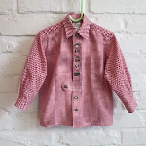 May include: Red and white checkered long-sleeve shirt with decorative buttons. The shirt has a classic collar and a small pocket detail. The shirt is hanging on a green hanger against a white brick wall.