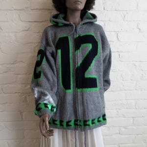 May include: A gray knit zip-up hoodie featuring a large black number 12 on the front with bright green trim. The garment includes a hood with green interior detailing, long sleeves with green accents at the cuffs, and a green patterned hemline.