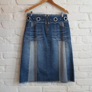 May include: A blue denim skirt with a knee-length hemline, hanging on a wooden hanger. The skirt features a front zip, pockets, and decorative metal eyelets along the waistband. The skirt has a two-tone design with a lighter denim panel.