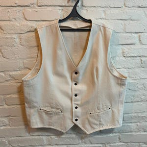 Puede incluir: Chaleco color crema con cuello en V y botones negros. El chaleco tiene dos bolsillos delanteros y está colgado de una percha negra. El fondo es una pared de ladrillos blancos. Una prenda clásica.
