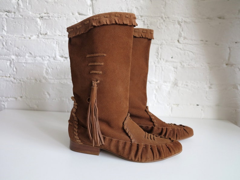 May include: A pair of brown suede boots with a knee-high design. The boots feature fringe detailing at the top, decorative stitching, and a tassel. The soles are a darker brown, and the boots have a low heel.