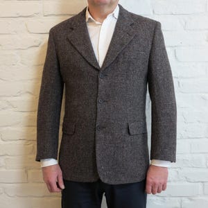 Peut inclure: Un blazer en tweed marron avec une fermeture à trois boutons, deux poches avant et un revers cranté. Le blazer est porté sur une chemise blanche. La veste est de style classique.