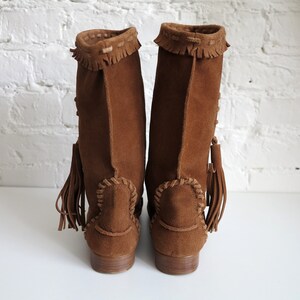 May include: A pair of brown suede boots with fringe and tassel details. The boots have a low heel and decorative stitching around the ankle and top of the boot. The boots are displayed against a white brick wall.