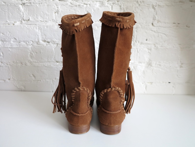 May include: A pair of brown suede boots with fringe and tassel details. The boots have a low heel and decorative stitching around the ankle and top of the boot. The boots are displayed against a white brick wall.