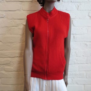 May include: A bright red sleeveless vest with a full-length zipper and a high collar. The vest has ribbed detailing on the shoulders and a pocket on each side. The bottom hem is ribbed. The vest is displayed on a mannequin.