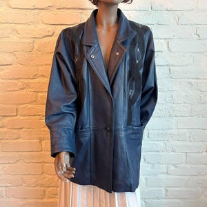 May include: A dark blue leather blazer featuring a notched lapel, long sleeves with wide cuffs, and a single button closure. The garment includes black textured panels along the chest and shoulders, with two front pockets at the waist.